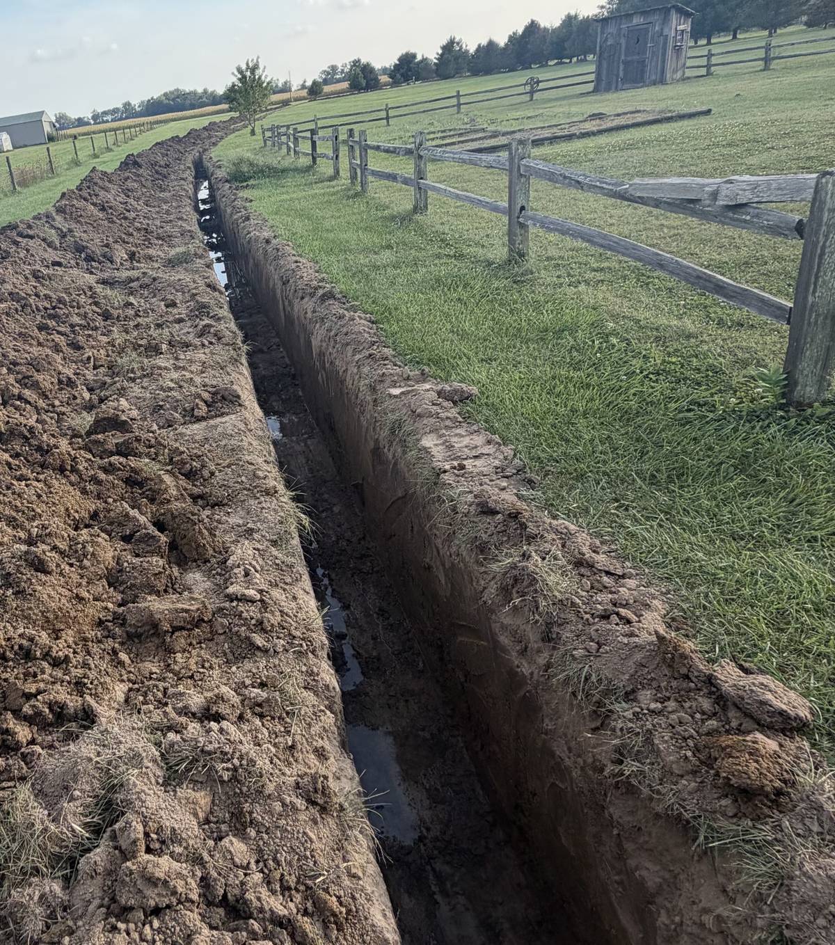 A long open trench cut along a split rail fence on a rural property